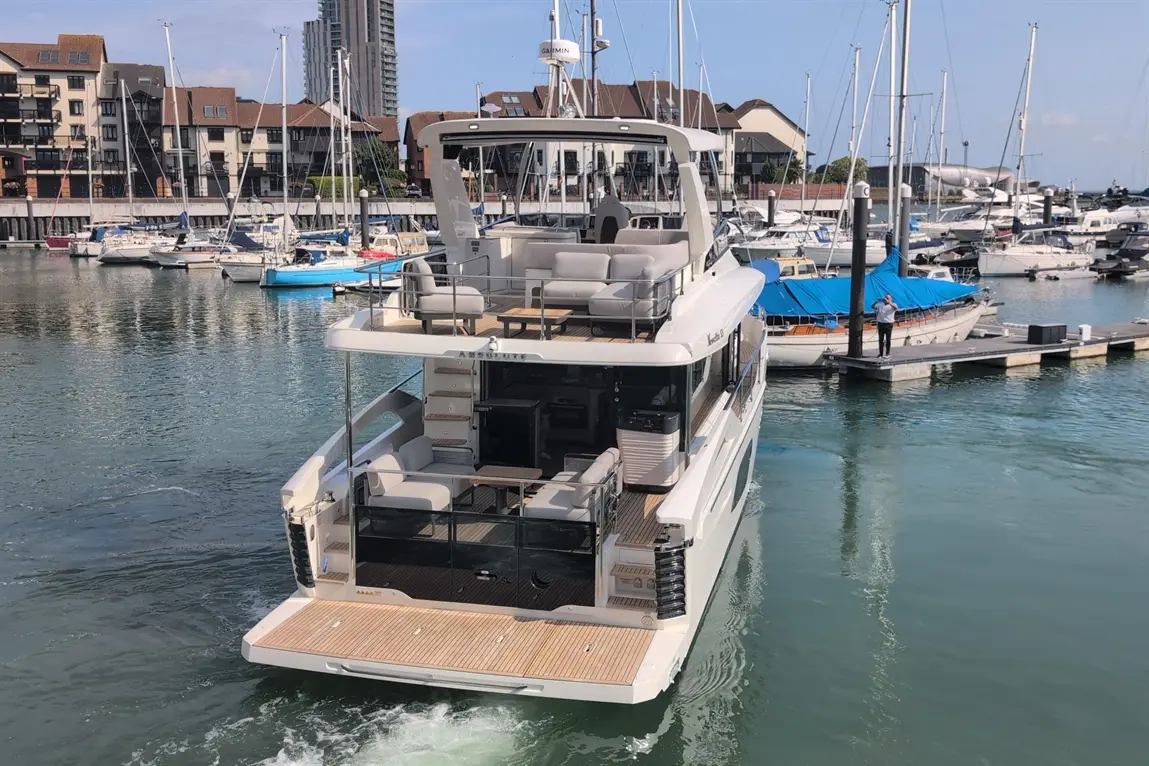 Luxury motor yacht at a marina, featuring outdoor seating and a person on the dock beside various boats and buildings.